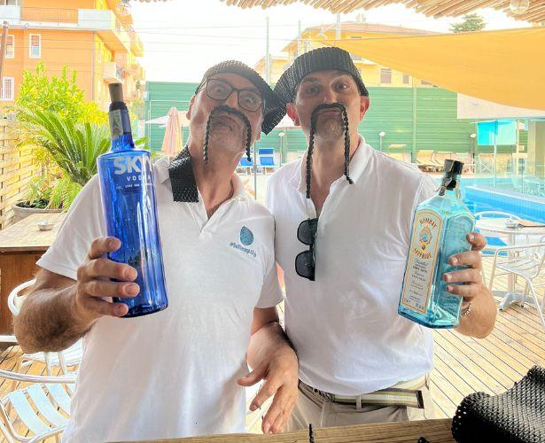 Due uomini con cappelli e bottiglie, sorridono in un ambiente all'aperto.